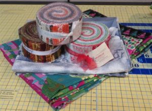 Three jelly rolls placed on top of two jelly roll race quilts that have been folded up as graphic for Jelly Roll Race, one of the June Special Events