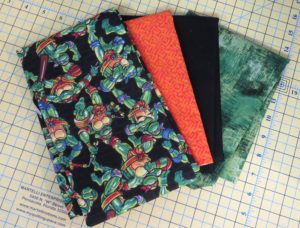 Photo of four fabrics used in square tuffet, feature fabric of Ninja Turtles, orange, black and green
